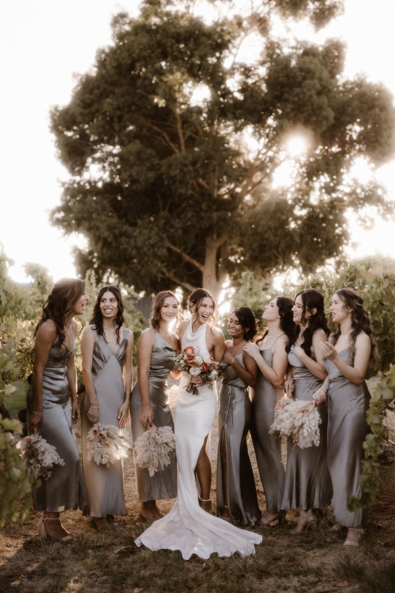 Bride in a high-neck gown with her bridesmaids in silver-grey dresses, laughing together at golden hour in a vineyard