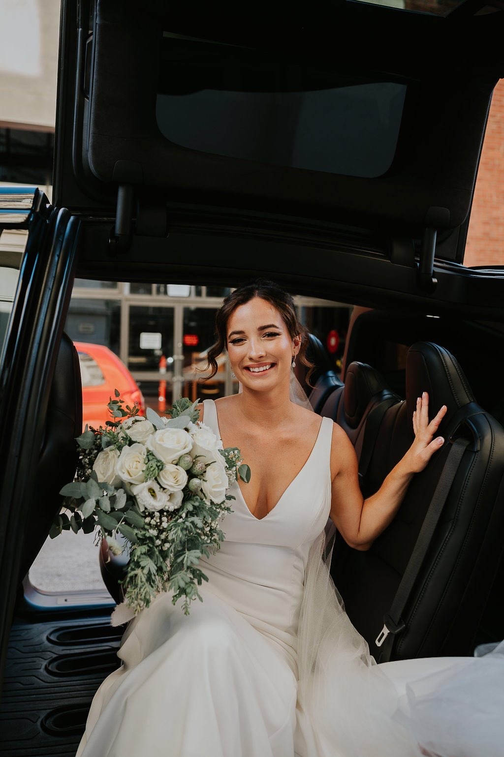 Real bride stepping from the wedding car with a soft brunette updo, face-framing tendrils and radiant glam makeup