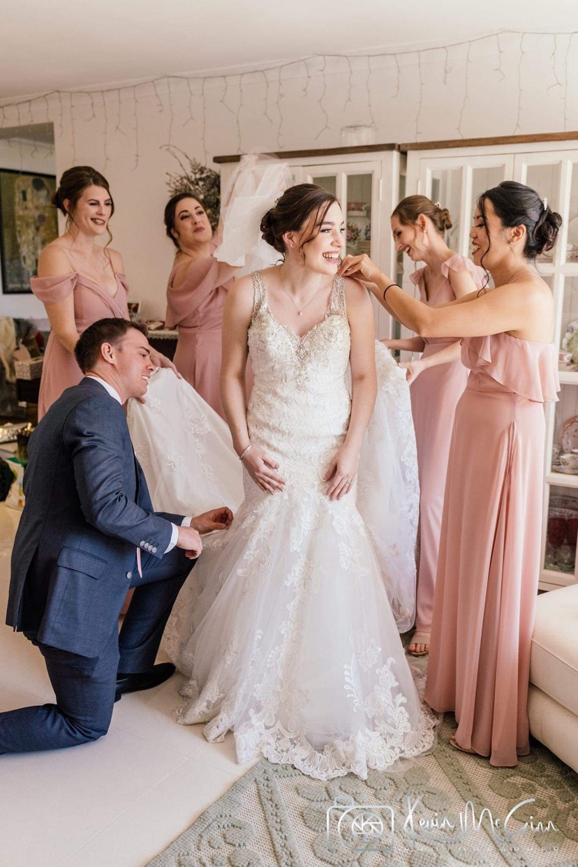 Real bride laughing as her bridal party helps her into her gown, polished brunette updo