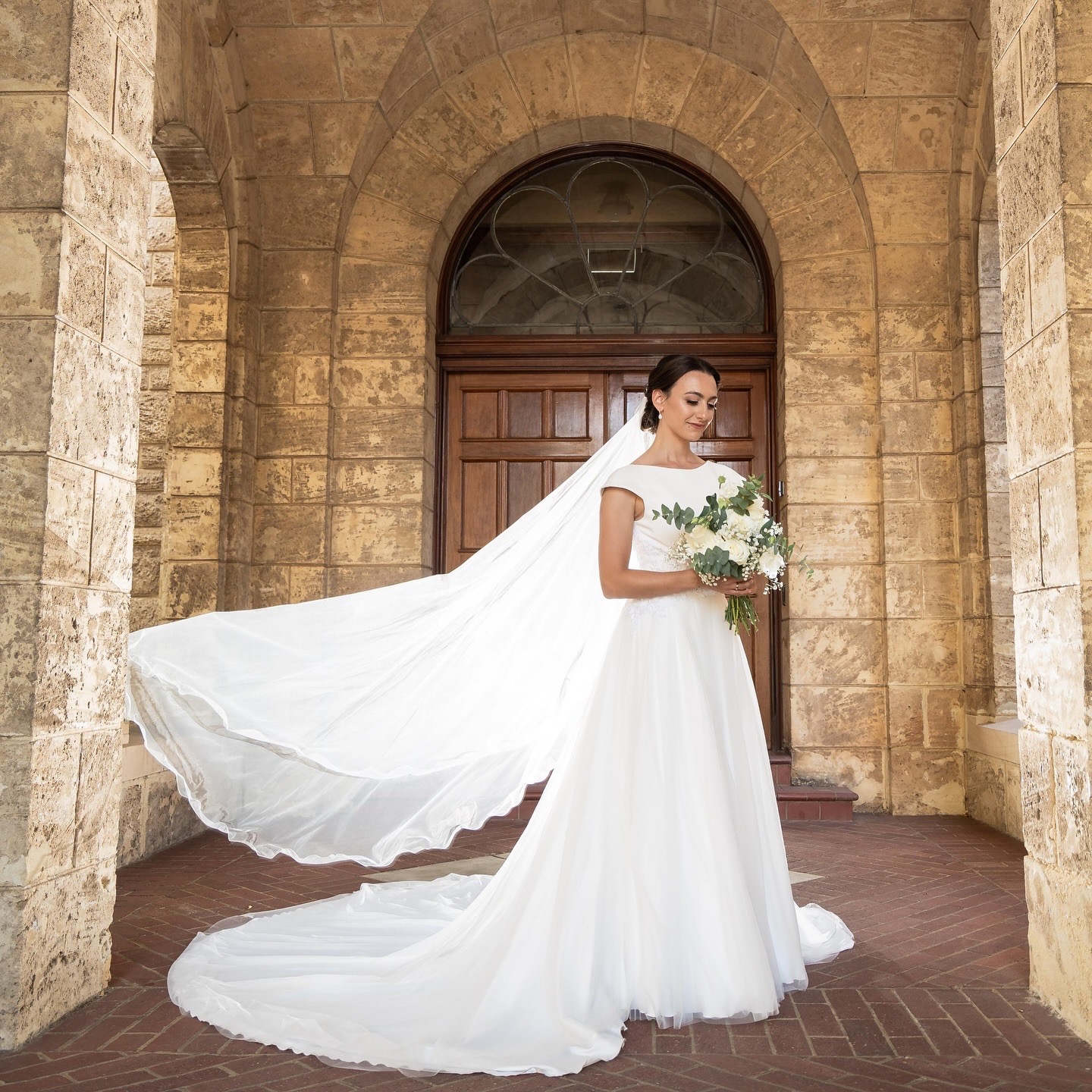 Real bride in a full tulle gown with a flowing cathedral veil and polished low bun in a stone archway