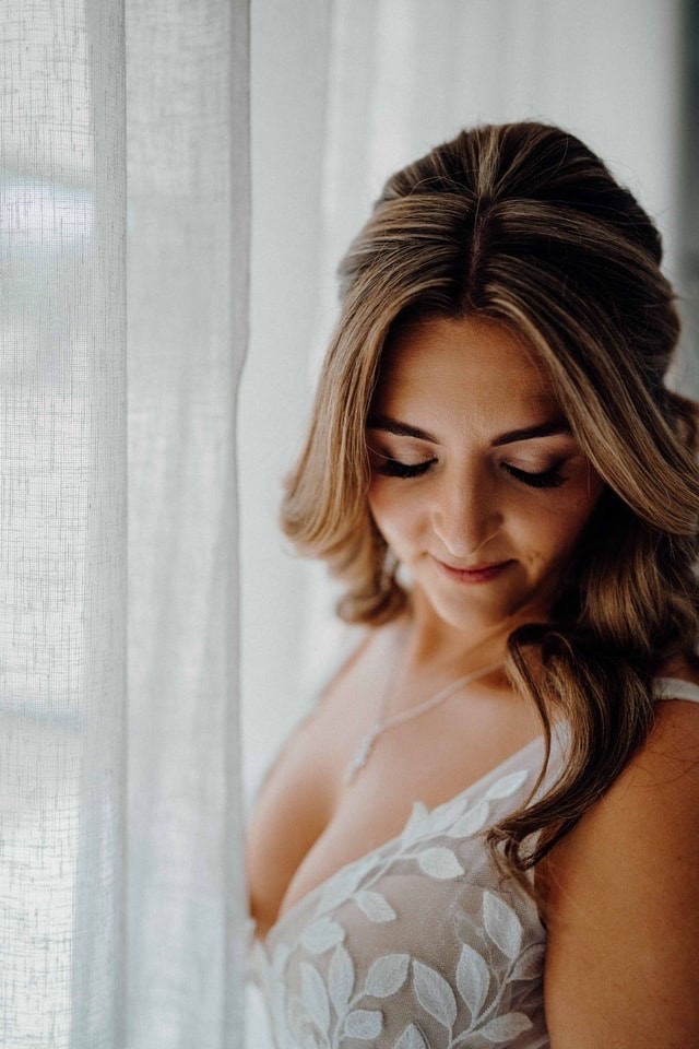 Real bride by the window with soft blonde waves and warm neutral soft glam makeup