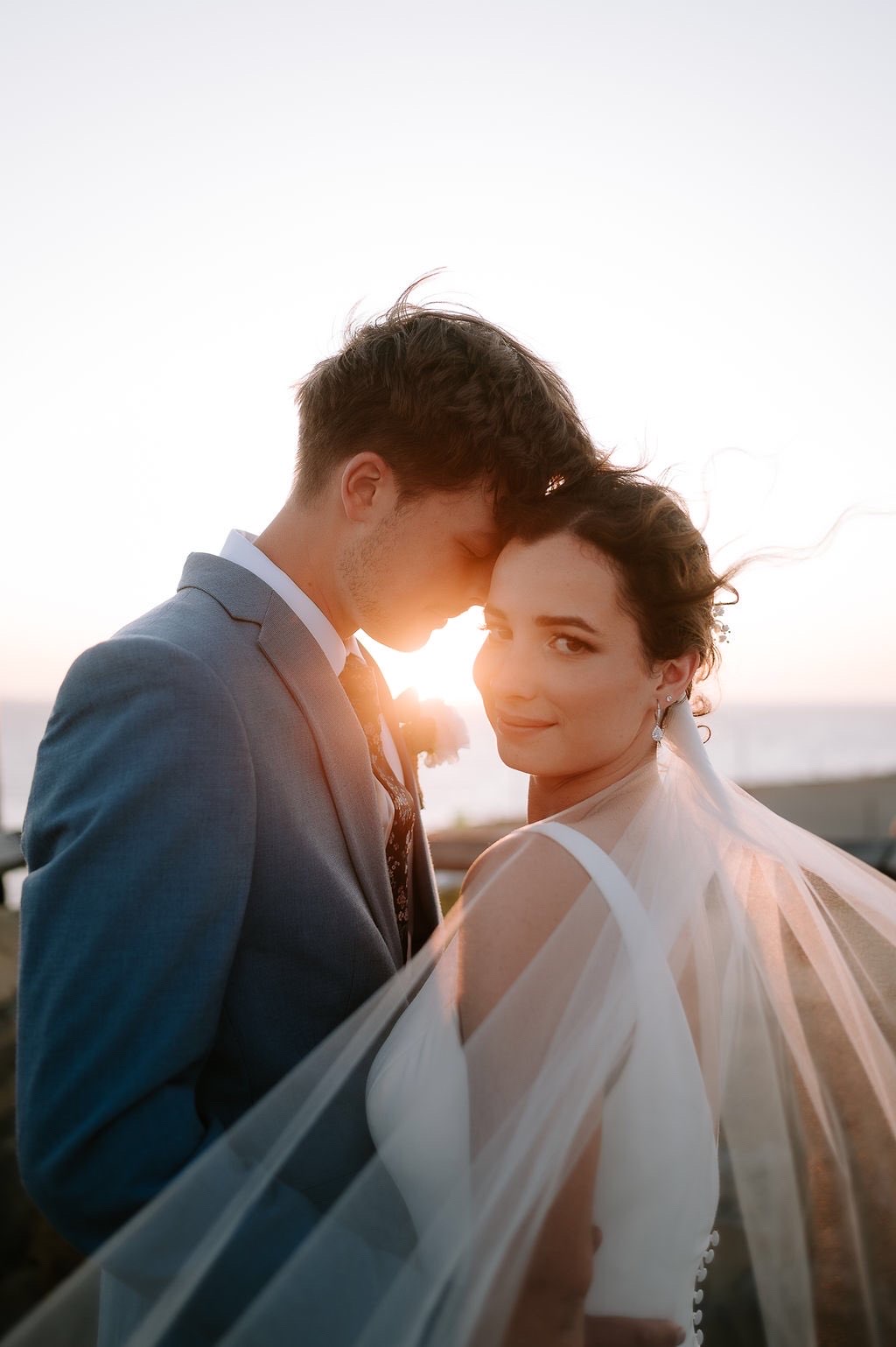 Real bride at sunset with a soft romantic updo, jewelled pin and floating veil, forehead to forehead with her groom
